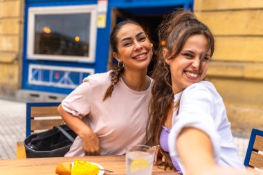 İki arkadaş bir kafede kahvaltı ederken selfie çekiyorlar. Seyahat maceraları sırasında bir neşe ve beraberlik anı yakalıyorlar.