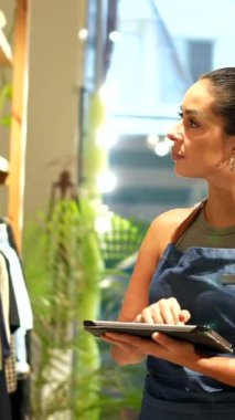 Genç profesyonel Latin kadın mağaza envanterini dijital tablet kullanarak yönetiyor, kendine güvenen, odaklanmış bir yaklaşımla ürünleri etkili bir şekilde organize ediyor.