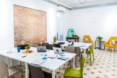 Parlak açık ofis alanı, dizüstü bilgisayarlı ve çizelgeli uzun beyaz masalar, dokulu tuğla bir duvar ve rahat sarı koltuklar ile rahat bir toplantı köşesinde