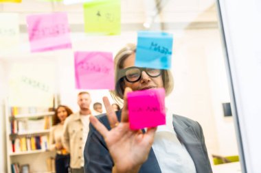 Olgun iş kadını cam bir tahtaya renkli yapışkan notlar yerleştirirken modern bir ofiste ekibiyle stratejik planlama oturumunu yönetiyor, işbirliği ve hedeflere odaklanıyor.