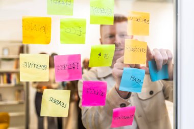 Modern ofis alanındaki bir adam cam duvarda çeşitli kavramlarla renkli yapışkan notlar düzenliyor ve stratejik bir planlama ve beyin fırtınası yapıyor.