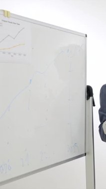 Kendine güvenen, gri saçlı üst düzey kadın yönetici, bir şirket toplantısında beyaz tahtadaki iş büyüme grafiğini açıklıyor, profesyonel bir ofiste liderlik ve stratejik planlama sergiliyor.