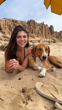 Kumsalda, kumlu bir plajda, mutlu kahverengi bir köpeğin yanında uzanmış gülümseyen genç bir kadın. İkisi de arka planda kayalık kayalıklar olan bir şemsiyenin altında dinleniyor.