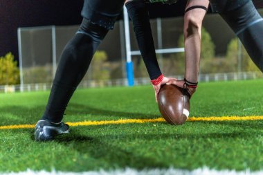 Koyu renk üniformalı Amerikalı futbolcu gece yeşil çimenliğe kahverengi bir top yerleştiriyor. Düşük açı başlangıç vuruşundan önce odak noktasını yakalıyor.