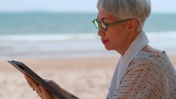坐在沙滩上使用平板电脑的亚洲老年妇女 — 图库照片