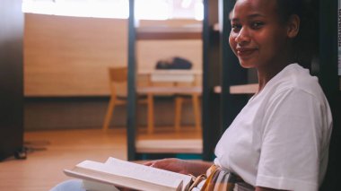 Kütüphanenin kitaplığının önünde oturan ve sınava hazırlanan bir kitap okuyan bir kız öğrenci..