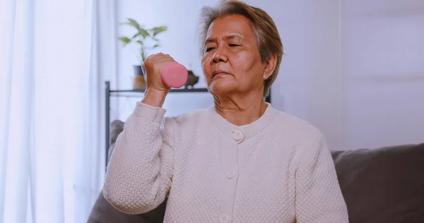 Asian old woman doing exercise with dumbbells at home. Elderly woman maintains her health by working out in every day.