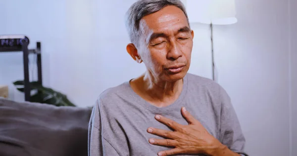 アジアの老人は自宅でソファーに座っている間呼吸が困難です ロイヤリティフリーのストック写真