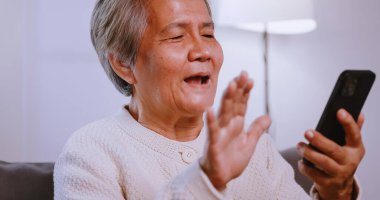 Mutlu Asyalı yaşlı kadın evde kanepede otururken akıllı telefon kullanıyor..