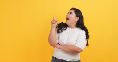 Beyaz tişört giyen tombul bir Asyalı kadın sarı arka plandaki bir şeyi işaret ediyor..