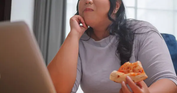 A chubby Asian woman enjoying pizza while relaxing in living room at home.