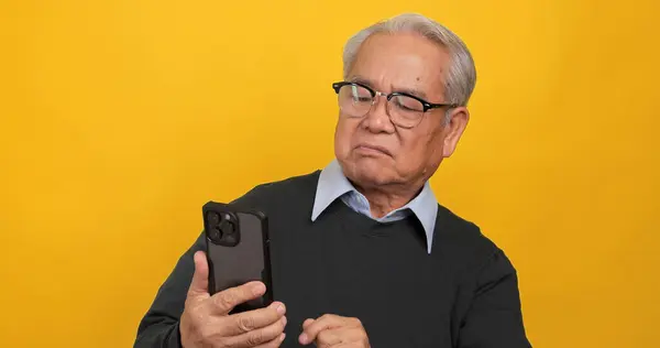 An old Asian man expressed his displeasure when he saw something on his phone. Isolated on yellow background in the studio.