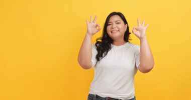 Beyaz tişört giyen tombul bir Asyalı kadın sarı arkaplandaki bir şeyle anlaşma ve tatmin jesti yapıyor..