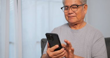 Asyalı yaşlı adam evdeki kanepede otururken akıllı telefon kullanıyor.