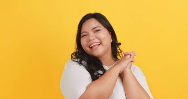 Beyaz tişört giyen tombul bir Asyalı kadın sarı arka planda kendine güvenini ve gururunu gösteriyor..