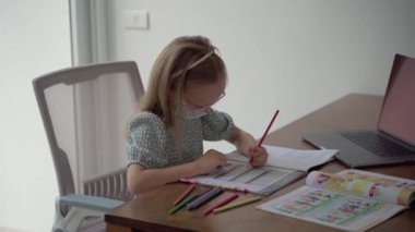 İlkokul çağındaki gözlüklü zeki kız koruyucu bir maskeyle İngilizce ödevi yapıyor. Çocukların ilköğretim kavramı. Çocuk evde ders izliyor. Salgın