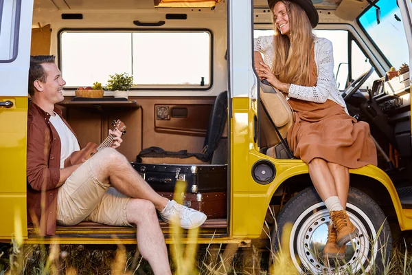 happy hipster couple sitting in camper van playing ukulele, making music together. attractive female in hat and handsome guy relaxing, having rest together, romantic time during adventure, travel