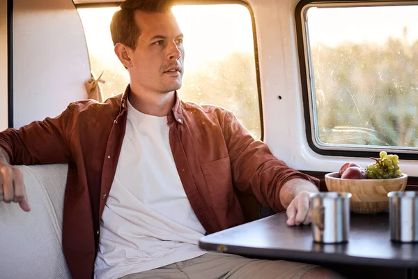 Side View On Calm Caucasian Handsome Male Sitting Behind Table In Mini Van, Looking At Side, Having Rest, in Contemplation, At Sunset. Traveller Tourist In Casual Wear. Trip Time