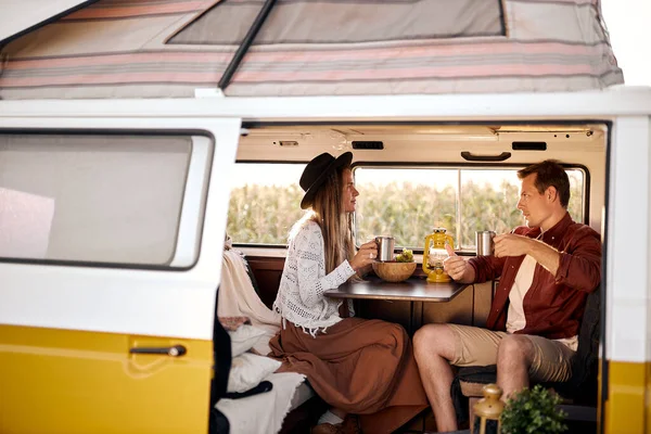 Lovely man and woman sit inside of van talking, drinking tea, discussing something during travel, trip together. caucasian couple in casual wear at adventure. people lifestyle, travel concept