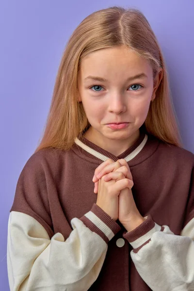 Forgive me. Little blonde teen kid child girl praying, looking at camera and making wish, asking with hopeful imploring expression, begging apology isolated on purple background. Children emotions