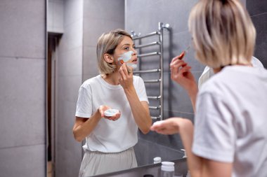 adorable female using skin face cream moisturizing lotion after taking bath. Pretty attractive lady wearing casual domestic clothes standing front of mirror in bathroom. Daily hygiene and skincare