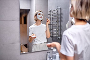 Beautiful young woman look in mirror applying cream on face in bathroom, pretty lady put facial moisturizer lifting hydrating moisturizing creme with brush, skin care treatment concept