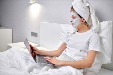 Sağlıklı bir kadın yüz maskesi takmış, dizüstü bilgisayar kullanıyor ve internette çalışıyor. Evdeki sıcacık zaman. Kişisel zaman ve spa konsepti. Coronavirüs salgını ve kendini karantinaya alma kavramı