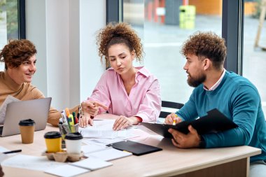 caucasian business team sit behind desk coworking, brainstorming. young curly man and woman in stylish clothes sit talking, holding papres documents. executive, success, people concept