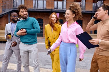 team of interracial business people take break outdoors, drink coffee after meeting, holding laptop and documents, leaving building, wearing stylish formal wear. african caucasian cooperation