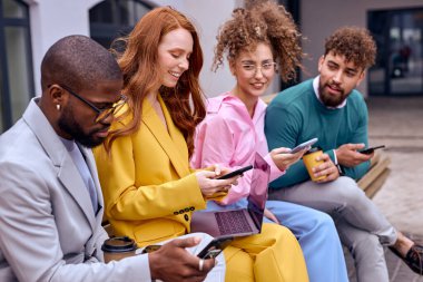 co-workers sit in row on bench using smartphone, dressed in stylish colourful formal clothes,afro and caucasian men and women after meeting, have rest during break. leisure. focus on redhead lady
