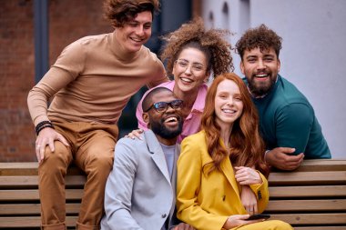 Excited successful group of people enjoying free time together outdoors. Men and women in stylish formal outfits after working in ecommerce business office, laughing,sitting on bench