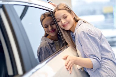 Cute Female Hugging New Car In Dealership Office, Overjoyed Customer Lady Leaning At Surface Of Luxury Automobile, Enjoying New White Vehicle. Smiling Caucasian Woman In Auto Salon