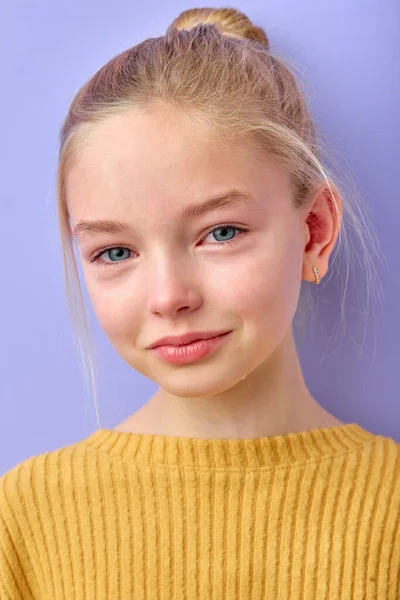Portrait of beautiful young blonde kid crying with tears smiling on camera in studio on purple background. Charming cute child in yellow shirt is stressed, sad and offended