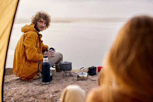 Handsome curly european guy in yellow coat sit on ground looking at girlfriend in tent, enjoying rest in nature, river in the background in the evening. thermos and small saucepan next to male.