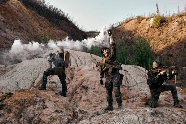 Three fearless Military Men call for help spraying artificial smoke standing on mountain, group of soldiers waiting for comrades during military operation outdoors, in uniform with rifles