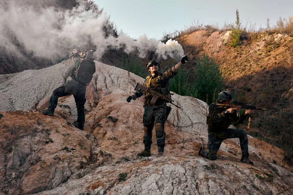 Three Military Men call for help spraying artificial smoke standing on mountain, group of soldiers waiting for comrades during military operation outdoors, in uniform with rifles