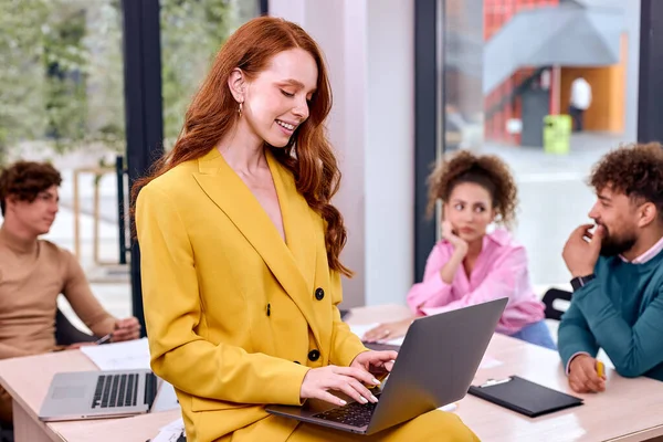 Pleasant Woman Working At Laptop During Meeting In The Background, Executives Gathered In Office, Discussing Plans And Ideas On Project. Redhead Beautiful Lady Sitting On Top Of Desk, Posing At Camera