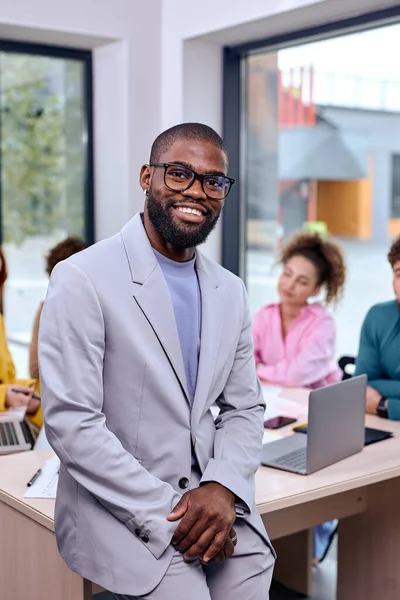 Portrait Of African American Man In Suit Standing In Busy Creative Office With Caucasian Colleagues In The Background. Smiling Successful Black Guy Posing At Camera. Business People Concept