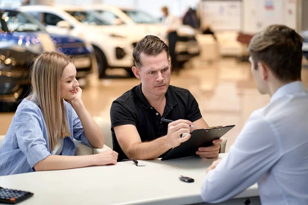 auto business, sale and people concept. happy couple with dealer buying car and signing documents in auto show or salon. automobile, vehicle, transport concept. clients sit behind table have talk