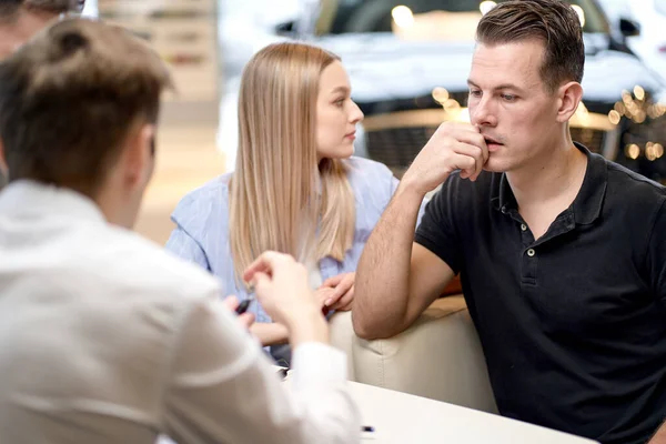 Young couple talking with car dealer in auto salon, man and woman listening to manager while sitting behind table in modern cars showroom. rear view on affable nice car dealer