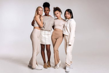 beauty portrait of diverse women, caucasian, asian and african american ladies with different skintone posing at camera, hugging each other, dressed casually in beige tone clothes. isolated