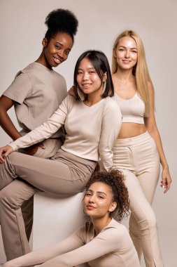 Excited smiling women with diverse appearance and natural make-up posing together, attractive ladies in casual clothes look at camera having cute smile on face, isolated on beige background