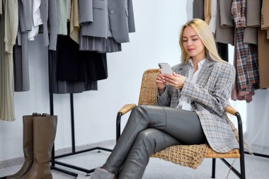 blonde stylist in casual gray clothes sit chatting with customer, online consulting, use smartphone, Women new collection of stylish clothes on hangers clothing rack rails, ashion design studio store