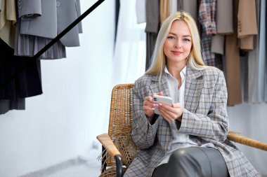 portrait of stylist in gray outfit sit chatting with customer, online consulting, use smartphone, Women new collection of stylish clothes on hangers clothing rack rails, ashion design studio store