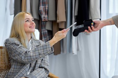Shopping App. Happy Stylish Caucasian European Blonde Female With Smartphone Buying Clothes Using Mobile Application On Phone Standing Near Clothing Rack In Cozy Showroom Studio, Smiling.