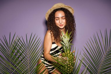 Happy young sexy woman in swimwear and straw brim hat posing isolated on purple color wall background studio. Summer hotel pool sea rest sun tan concept. Curly caucasian lady with pineapple