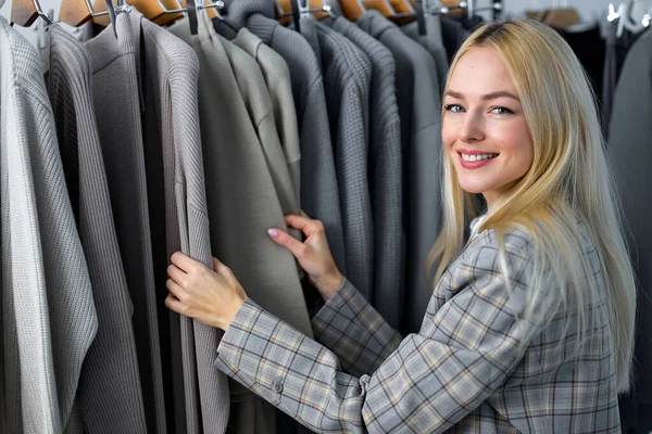 Successful young woman. Clothes boutique business. Personal fashion stylist. Attractive caucasian lady with trendy clothes on rack, look at camera. In cozy modern showroom shop. fashion, design
