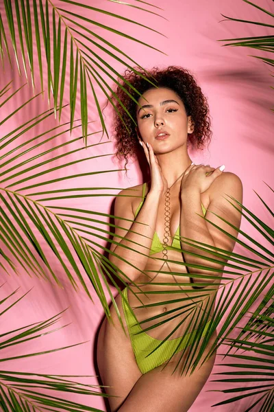 lovely lady on exotic resort having good mood relaxing resting in swim wear isolated pink color studio background. curly caucasian female with fit body in green swimwear with tropical exotic plants