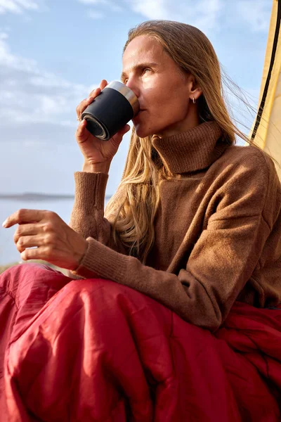 camper female sitting inside of tent and drinking hot coffee from thermos cup while spending time in nature, attractive caucasian lady enjoy the view landscape, feel good. travel concept