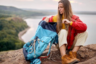 Confident caucasian female traveller unpacking backpack, river in the background, travelling alone, trekking and hiking. discovery, adventure concept. beautiful long haired woman in red coat